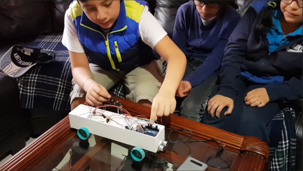 Three students from the Mexico demonstrate their robotic bus on the table in front of them
