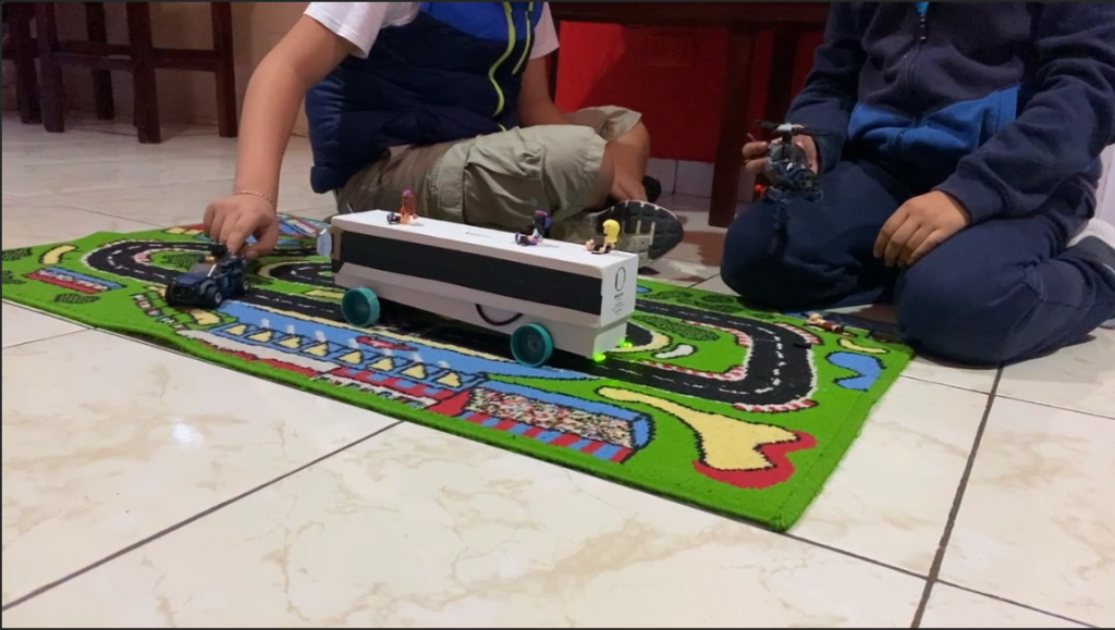 The robotic bus is on a play mat as the students show it's function to sense blockages in the road
